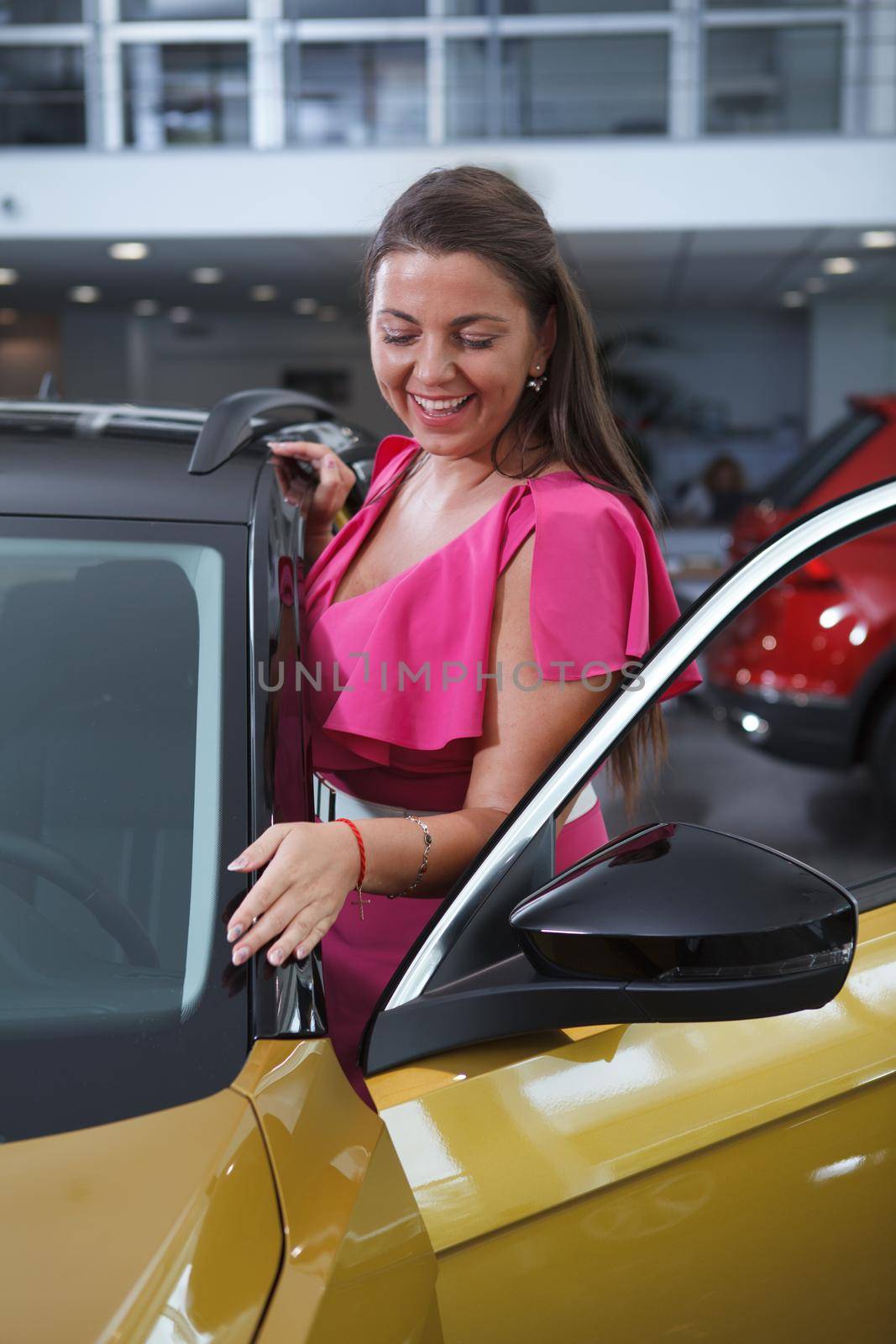 Vertical portrait of a cheerful beautiful woman looking at her new car at dealership