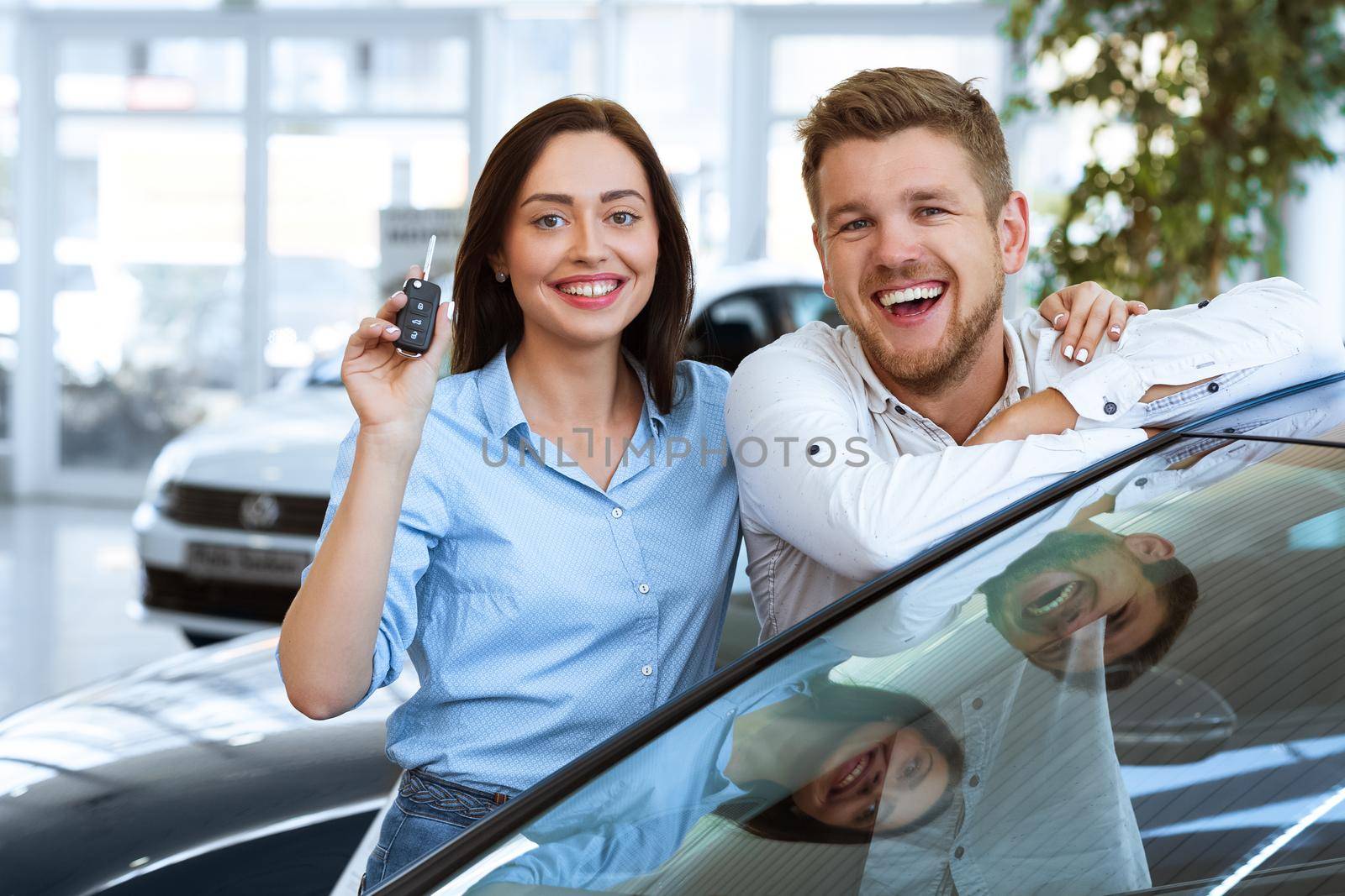 Our first car. Beautiful happy woman smiling showing the keys to a new car they just bought with her husband happy couple smiling to the camera at the dealership