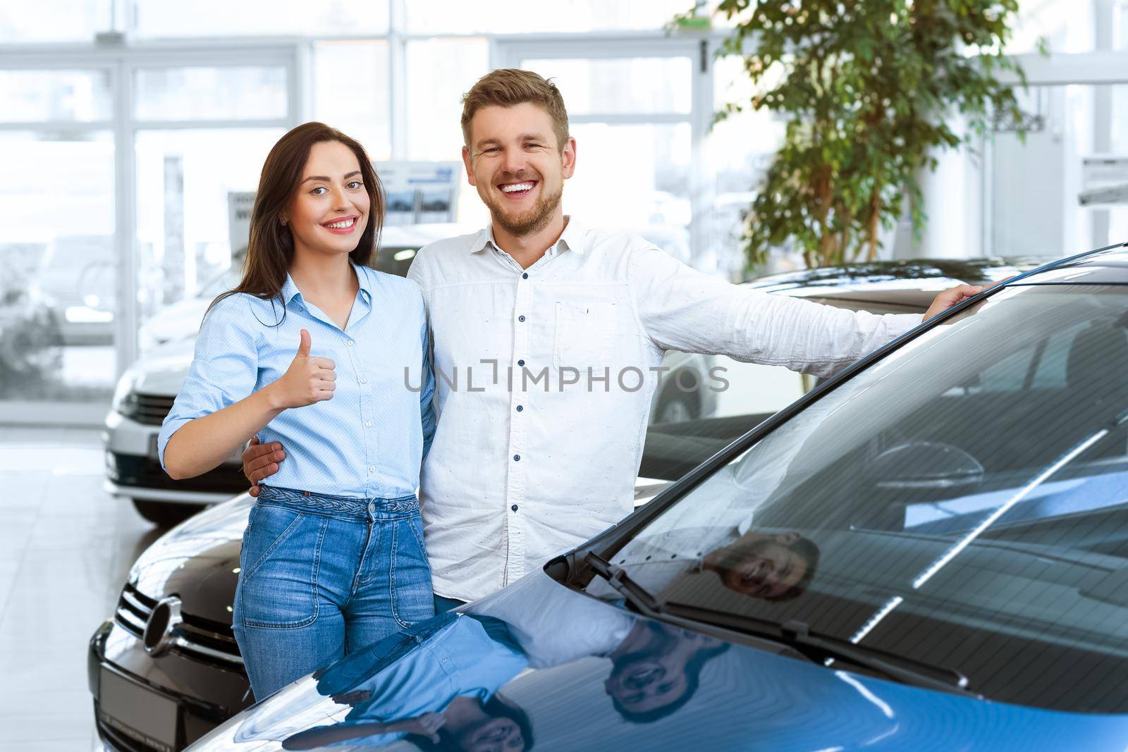 Buying a dream car. Portrait of a beautiful young couple buying a new car at the dealership handsome man laughing and hugging his wife while woman is showing thumbs up
