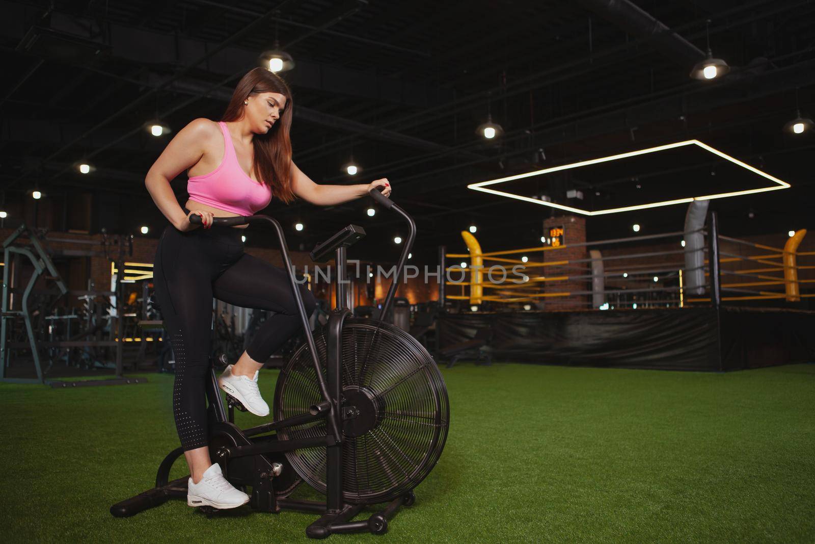 Beautiful focused young plus size woman exercising on air bike. Charming healthy plus size curvy woman cycling on air bike during cardio training, copy space
