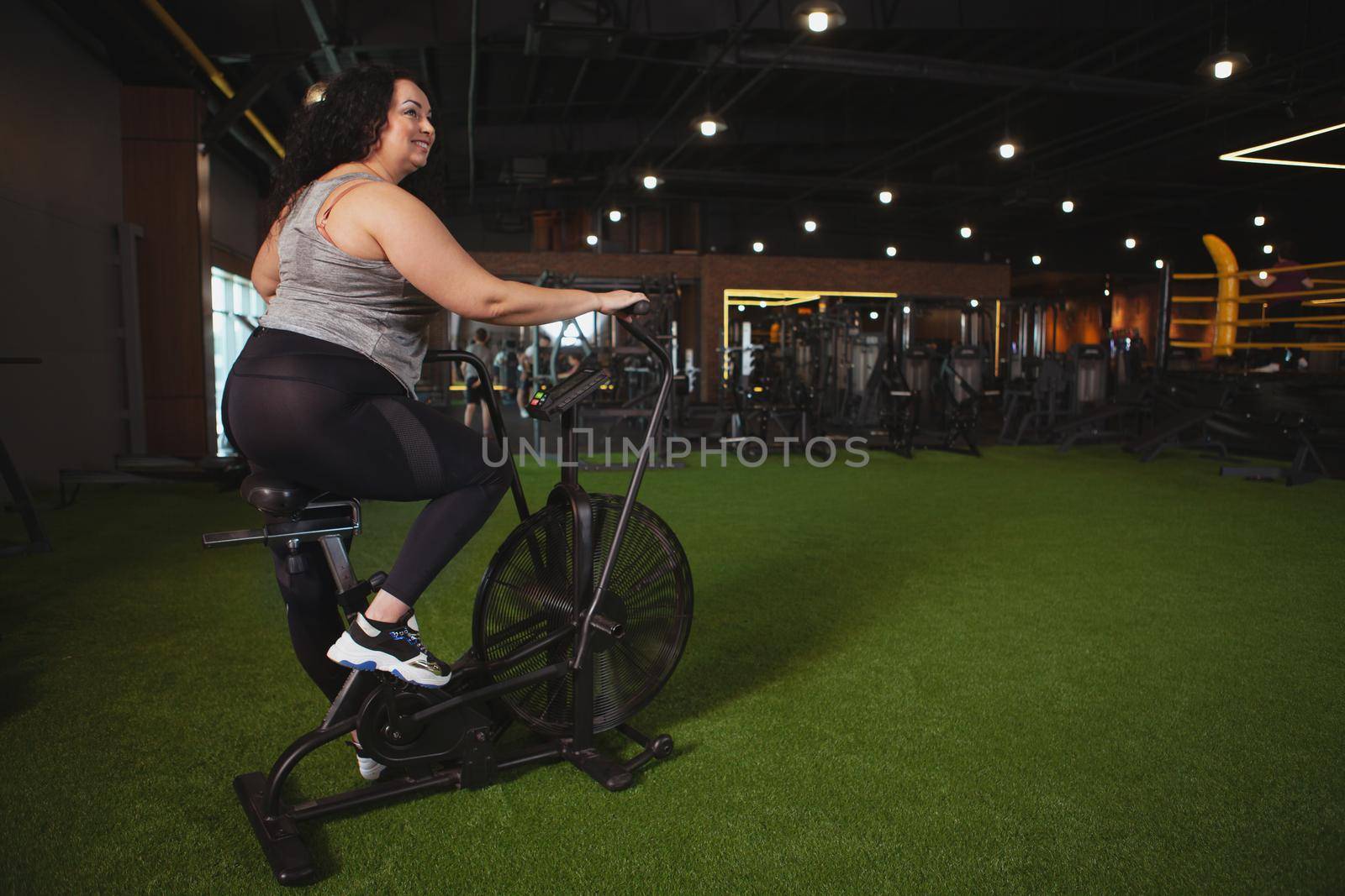 Rear view shot of a plus size sportswoman smiling, looking away, cycling on air bike at the gym, copy space. Cheerful overweight woman enjoying cardio workout at sport studio