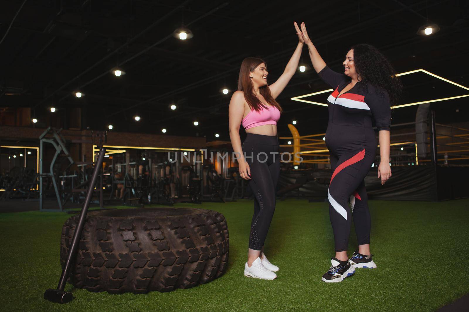 Two happy healthy plus size women laughing, giving each other high five after successful crossfit workout, copy space. Gym, fitness, body positive concept