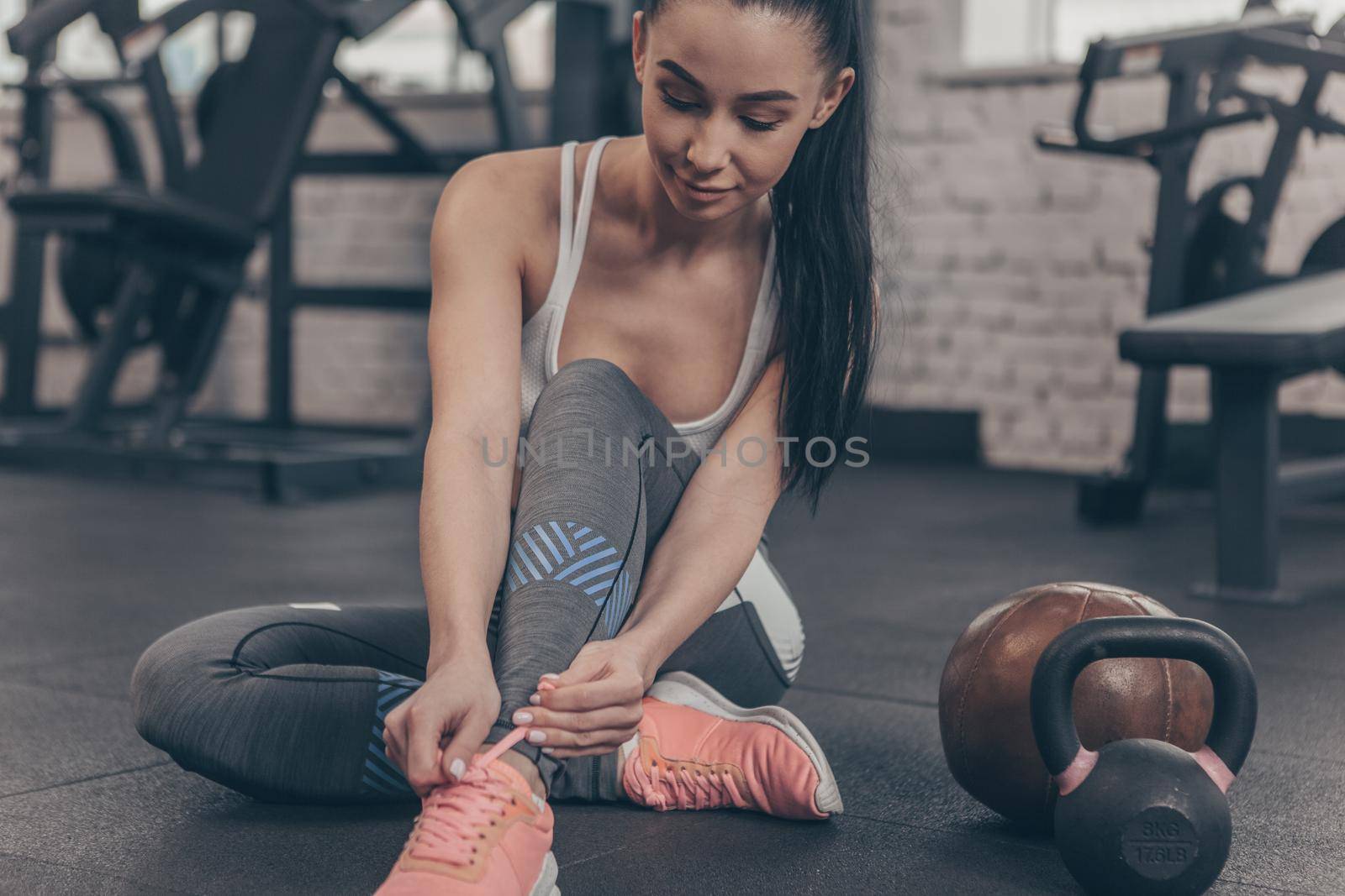 Cropped shot of a lovely young sportswoman tying her shoelaces, sitting on the gym floor, copy space. Beautiful fitness woman resting after working out at sports studio. Motivation, sportswear concept
