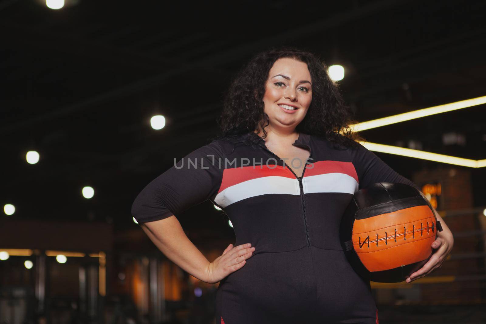 Happy beautiful mature overweight woman in sportswear smiling to the camera, holding medicine ball, copy space. Confident overweight female athlete posing proudly at the gym
