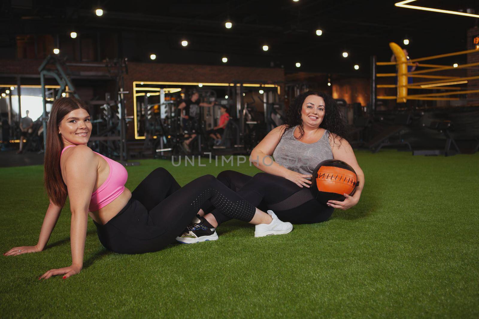 Lovely plus size young sportswoman smiling to the camera over her shoulder, enjoying working out with her friend at the gym, copy space. two happy women resting after exercising