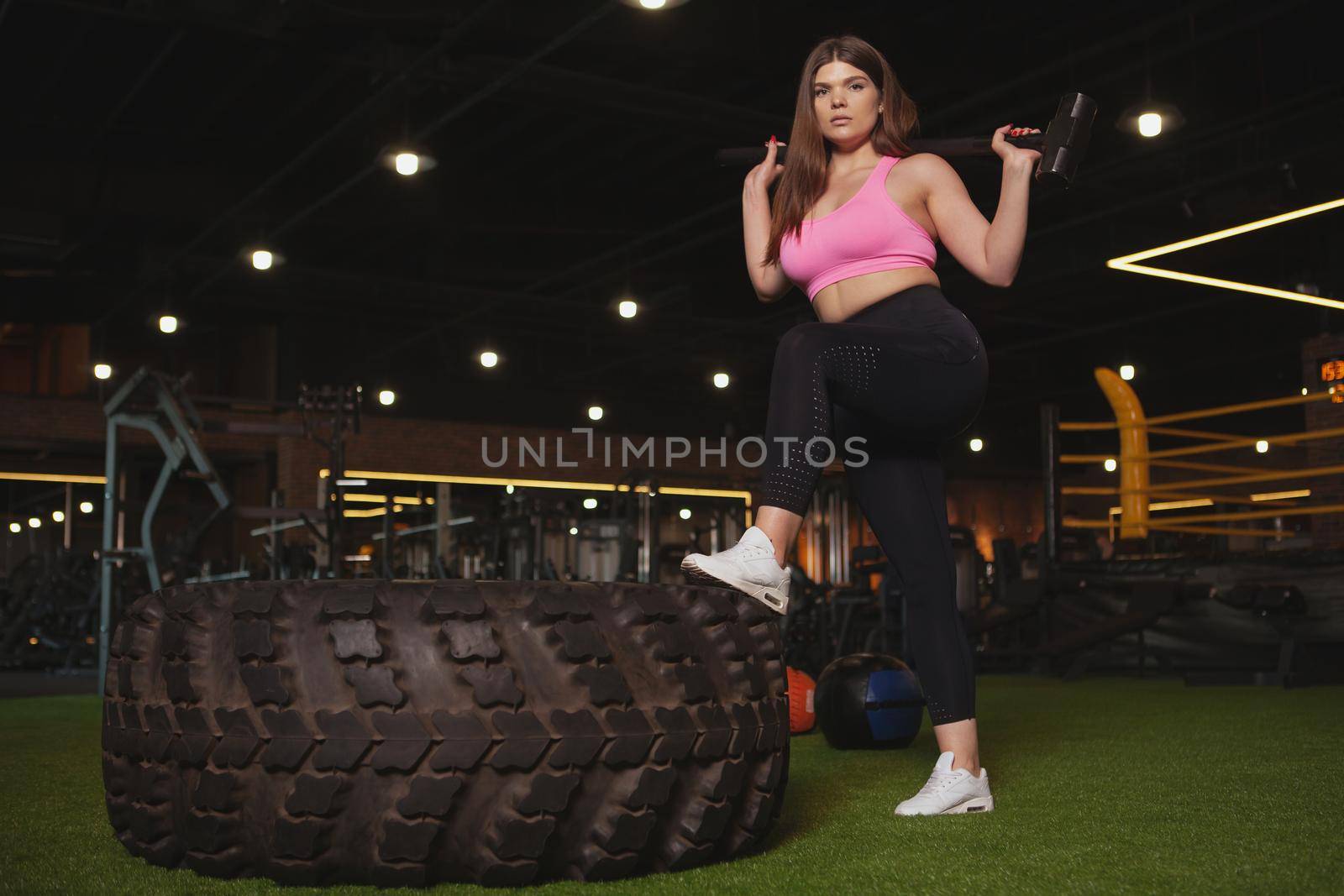Low angle shot of a confident beautiful plus size sportswoman looking fiercely to the camera, posing with a sledgehammer near heavy wheel at the gym, copy space