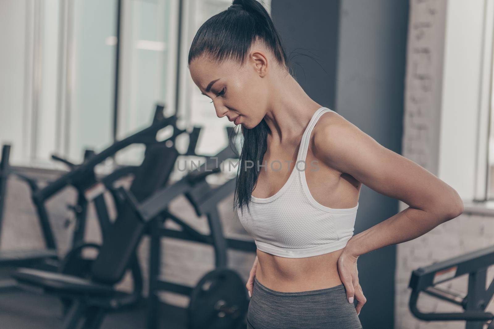 Beautiful athletic woman having backache after workout at the gym. Attractive fitness female having back or spine injury. Sportswoman rubbing her back, after tiring exercising. Injury, relief concept