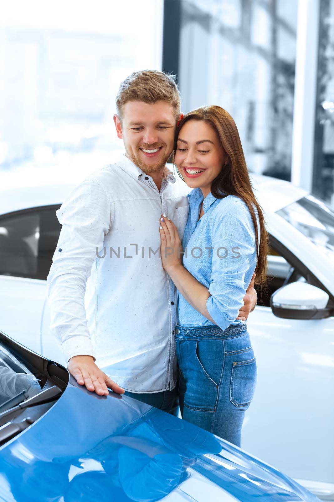 They dreamed of this day. Vertical shot of a handsome man hugging his happy girlfriend smiling cheerfully while touching a new car at the dealership