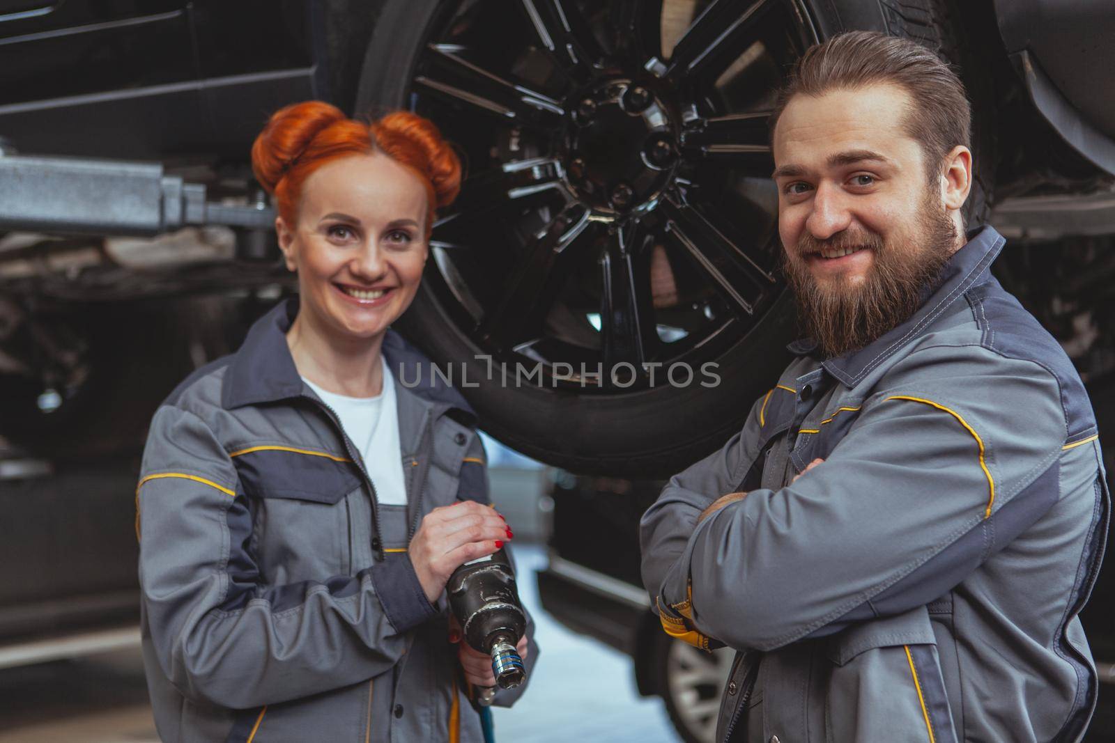 Cheerful beautiful female mechanic and her male colleague smiling to the camera, working at their car service station. Experienced car technicians repairing vehicles at the garage. Female equality concept