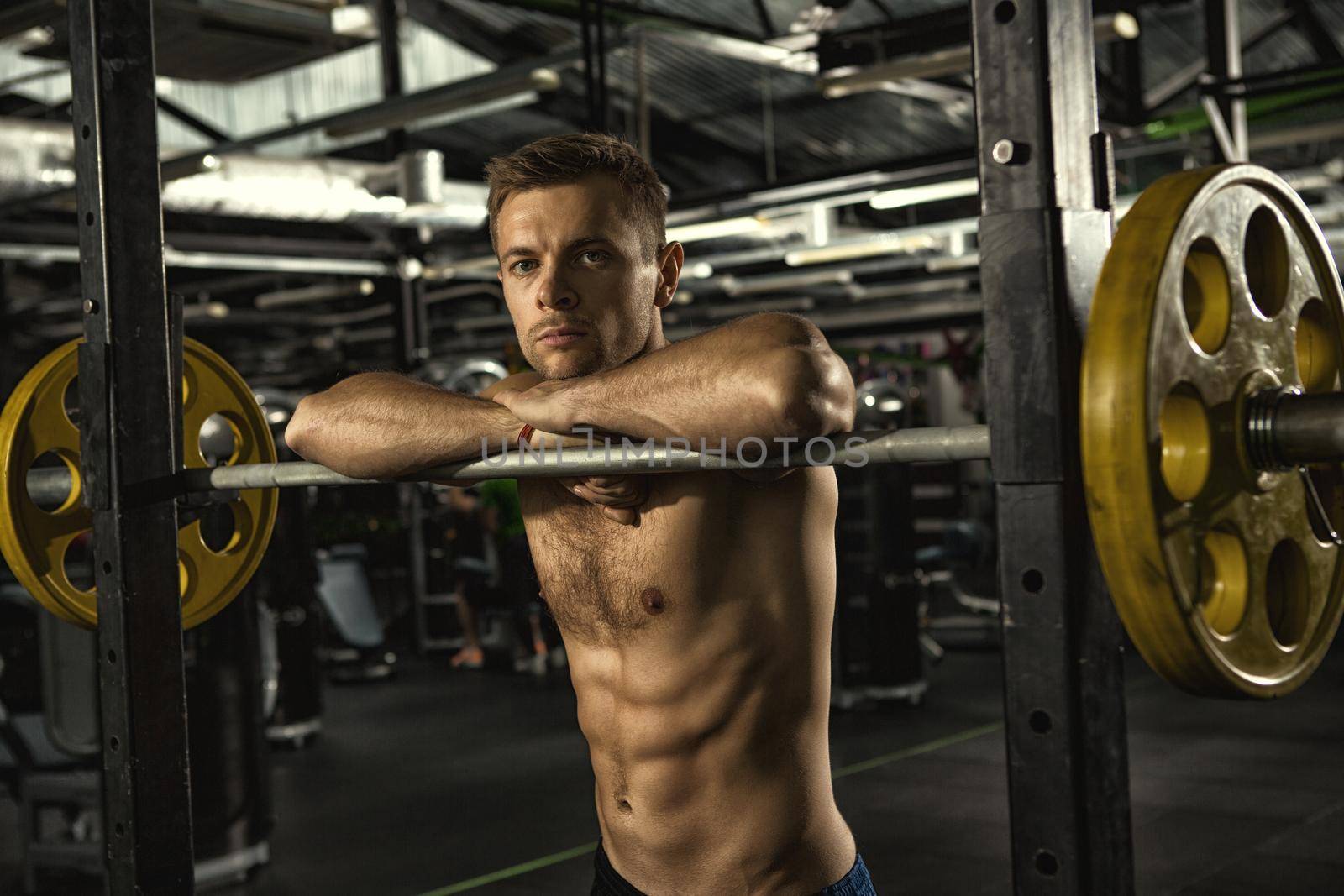 Resting in between sets. Young handsome shirtless sportsman looking to the camera leaning on the barbell resting after his workout at the gym exercising training fitness healthcare confidence concept
