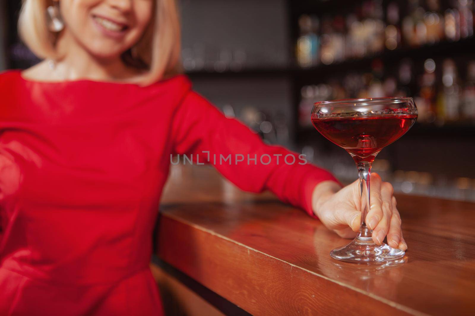 Close up of a cocktail glass in the hands of cheerful elegant woman in red dress. Woman drinking at the bar