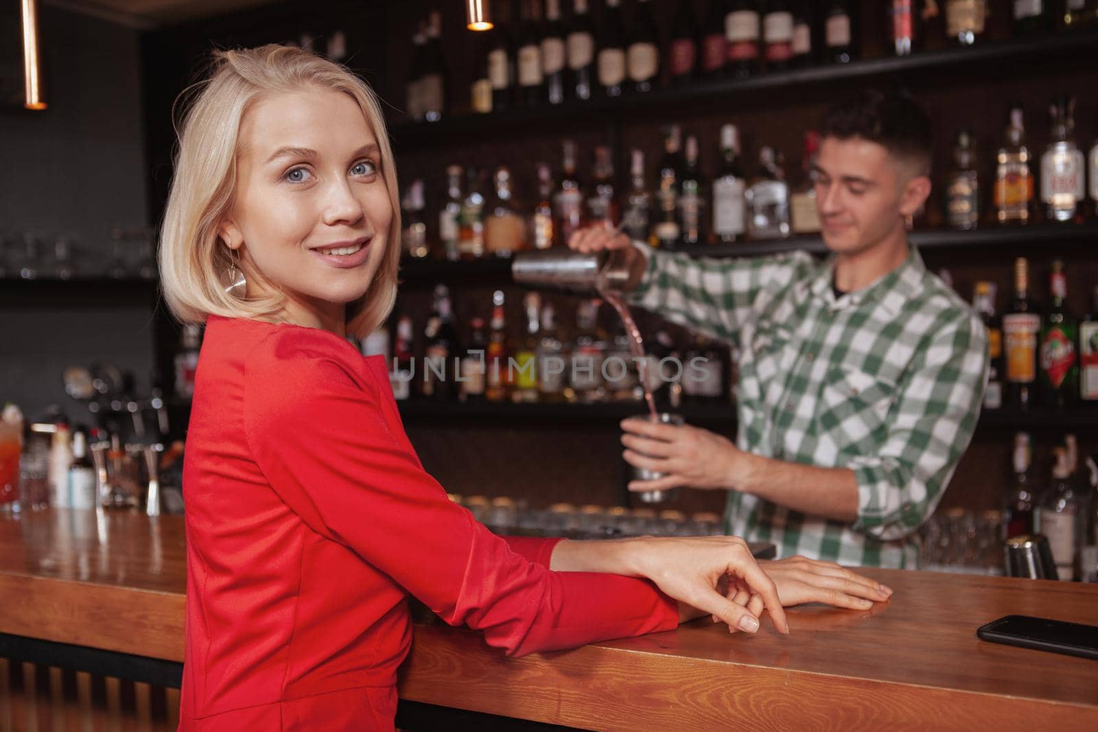 Beautiful happy blond woman smiling to the camera, waiting for her cocktail at the bar