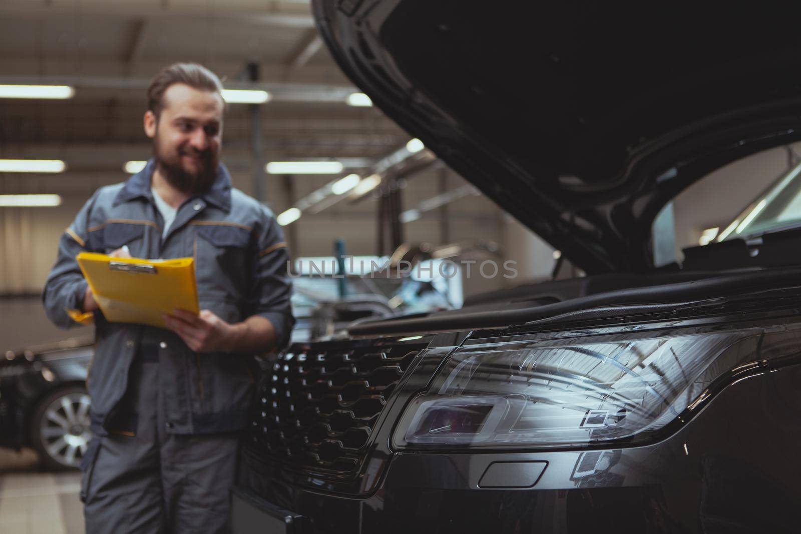 Selective focus on a car light, cheerful bearded male mechanic writing on his clipboard on the background. Professional car technician repairing cars at his garage, copy space