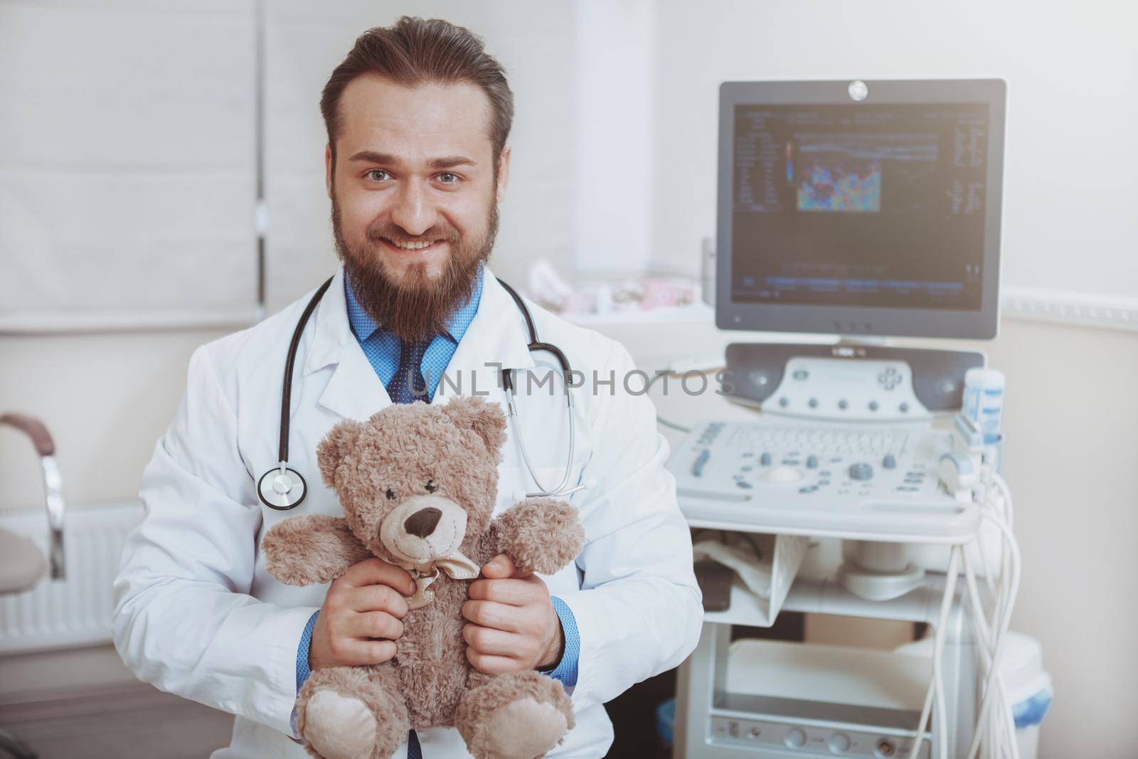 Happy male bearded doctor smiling to the camera, holding plush teddy bear toy, copy space. Cheerful pediatrician enjoying working at kids clinic. Pediatrics, childhood, health concept