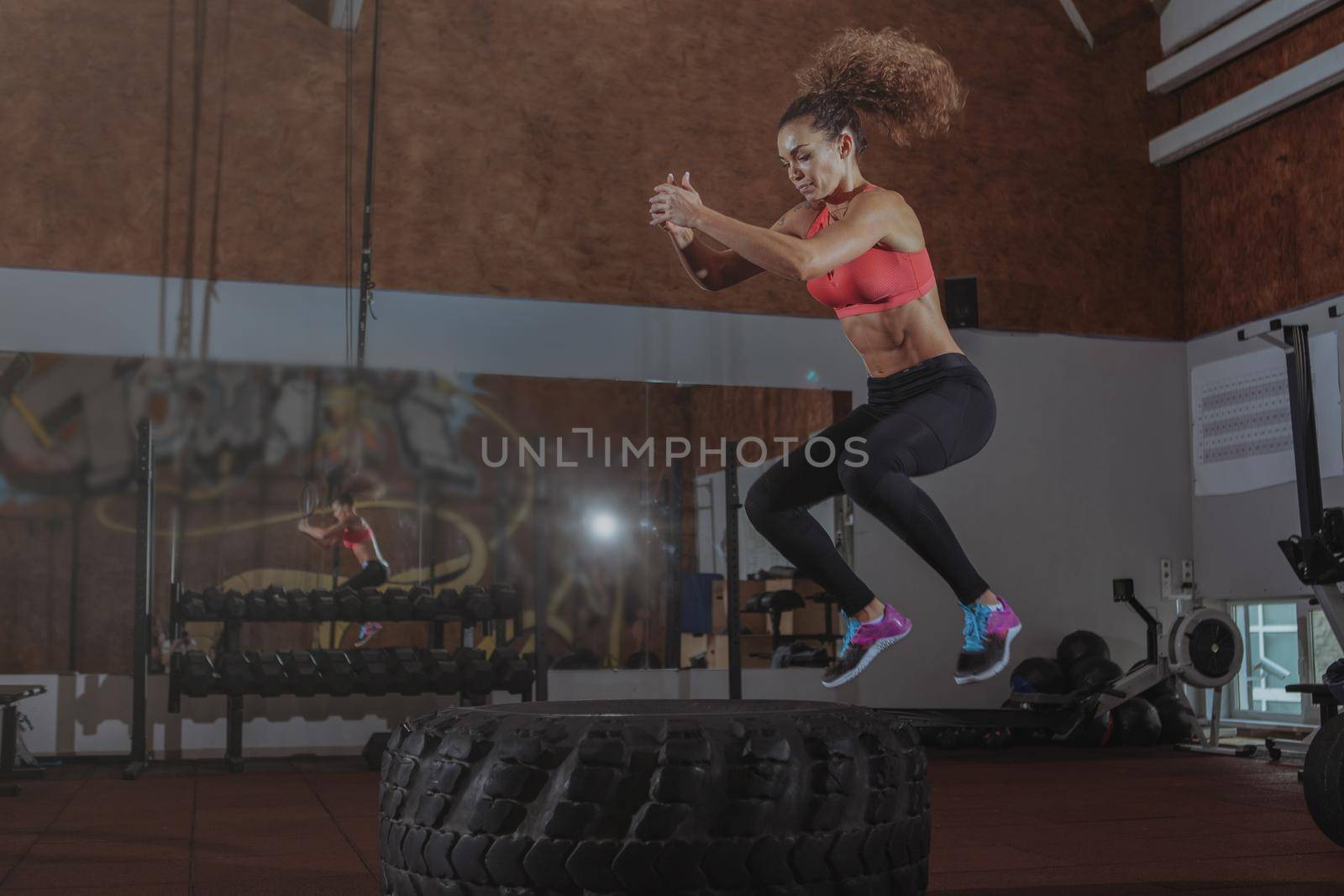 Beautiful fit sportswoman exercising at crossfit box gym, jumping high on a huge tire. Energectic female crossfit athlete working on her endurance and agility, copy space
