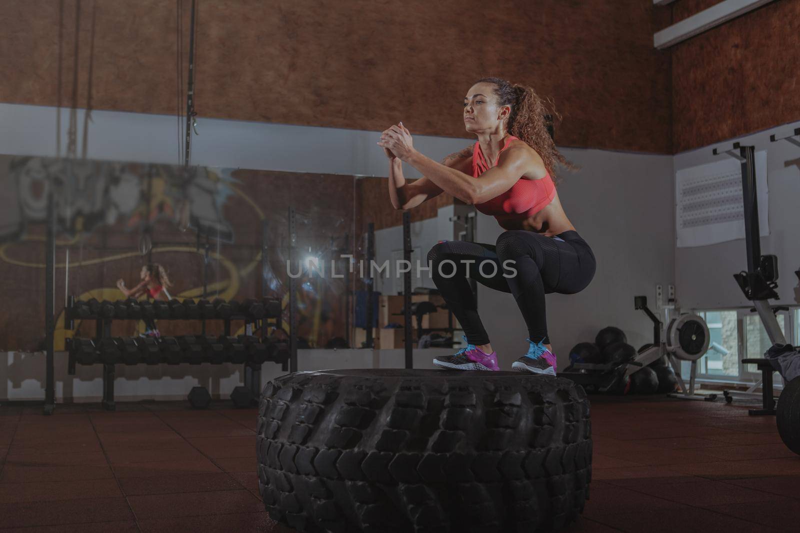 Attractive athletic woman jumping on a huge tire at crossfit box, copy space. Energetic female crossfit athlete working out at the gym, jumping high on a big wheel