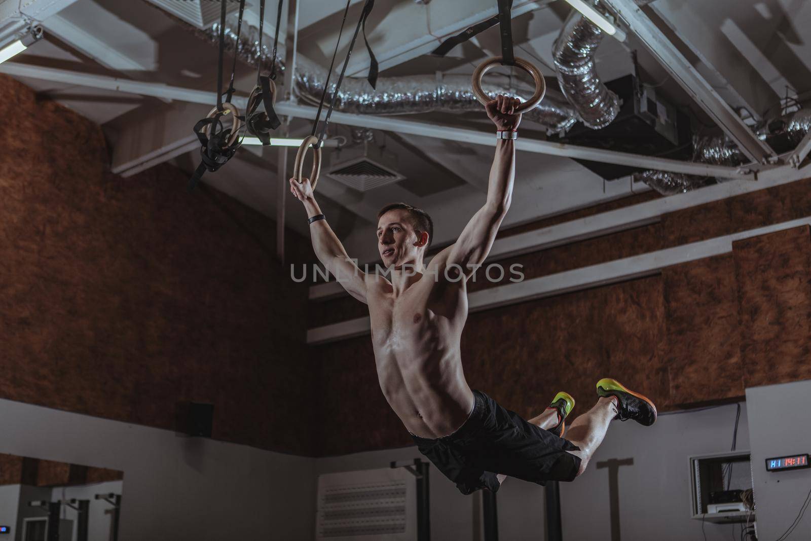 Low angle shot of a male crossfit athlete exercising on gymnastic rings. Shirtless strong sportsman working out on gymnastic rings at crossfit box gym