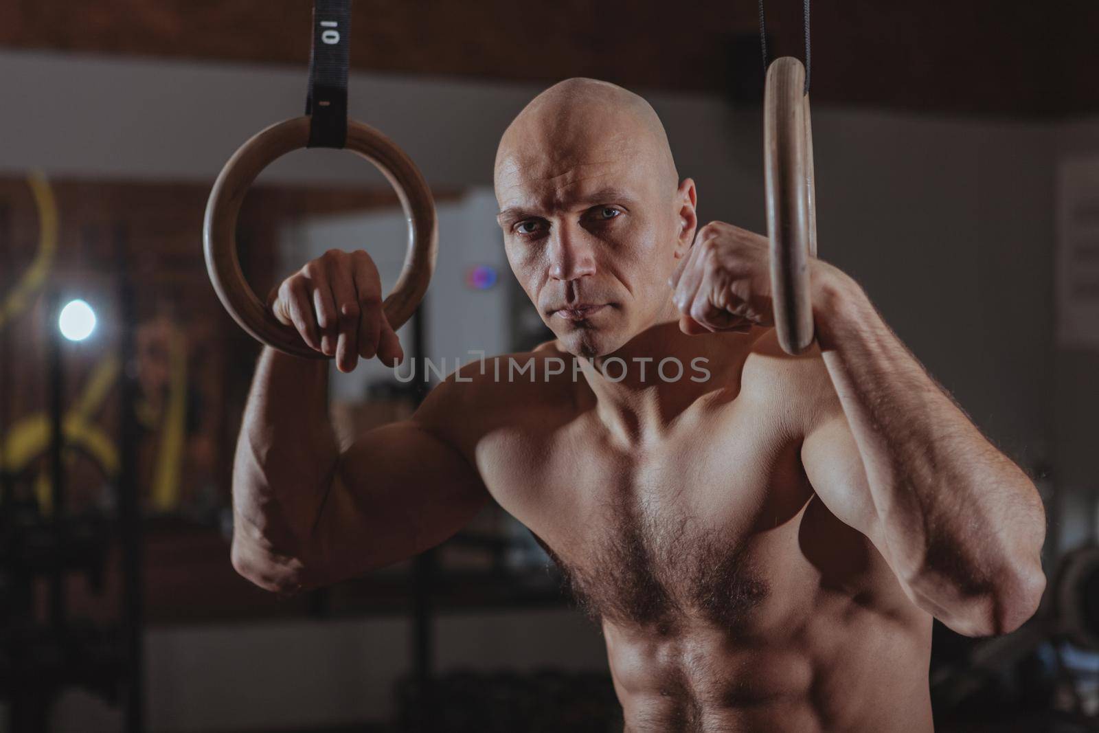 Close up of a shirtless mature crossfit male athlete holding gymnastic rings, relaxing at the gym after exercising. Muscular shirtless sportsman resting after workout