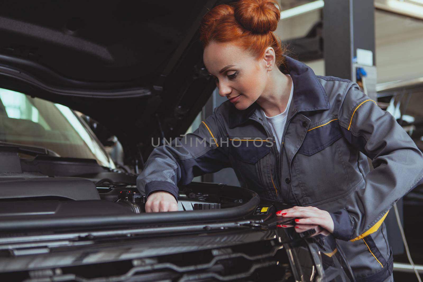 Experienced female mechanic servicing an automobile at her workshop. Lovely repairwoman working at the garage. Beautiful female technician in grey uniform working under the hood of modern SUV car, copy space