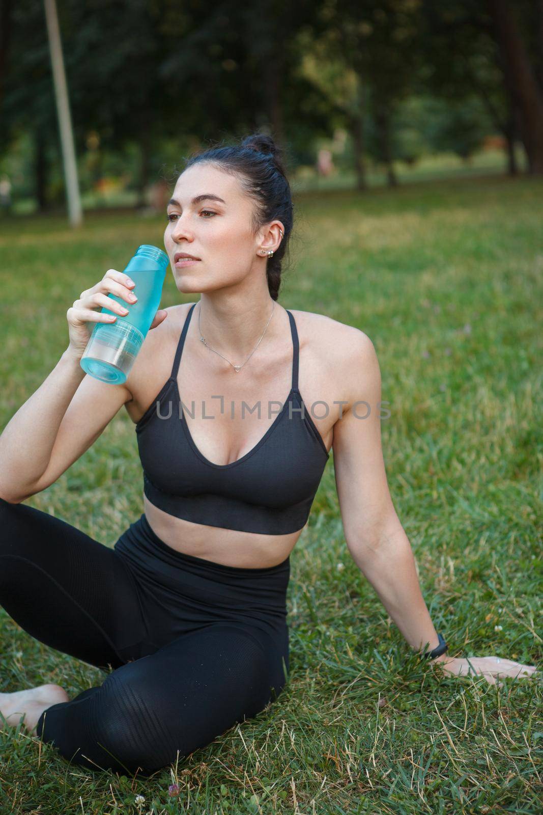 Vertical cropped shot of an attractive sthletic woman relaxing after outdoor workout, drinking water sitting on the grass