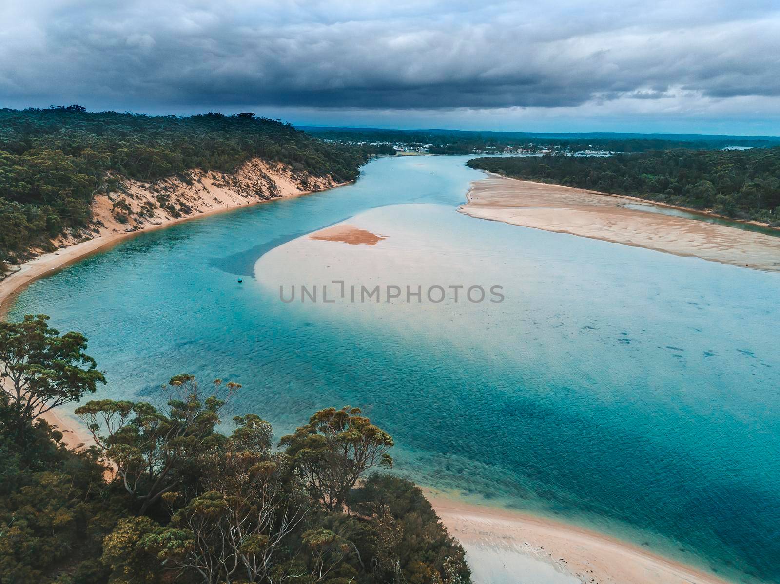 Storm clouds loom over the coastal tidal inlet with exposed sand bar.  
Australia