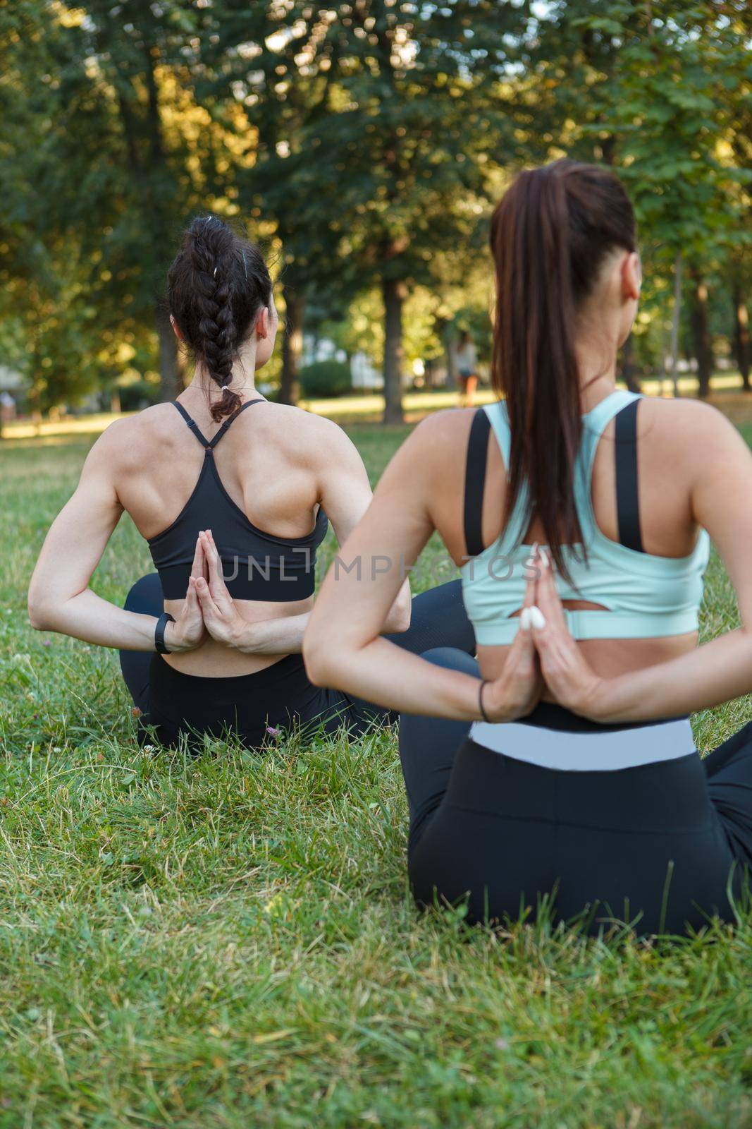 Rear view vertical shot of two women doing yoga outdoors, sitting in lotus positions on the grass