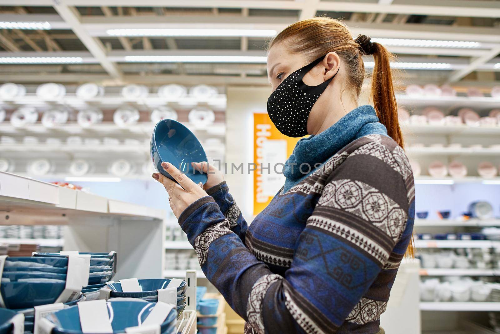 a woman in a protective mask chooses dishes and kitchen accessories in a supermarket, life with protection from infection in the new realities by vadiar