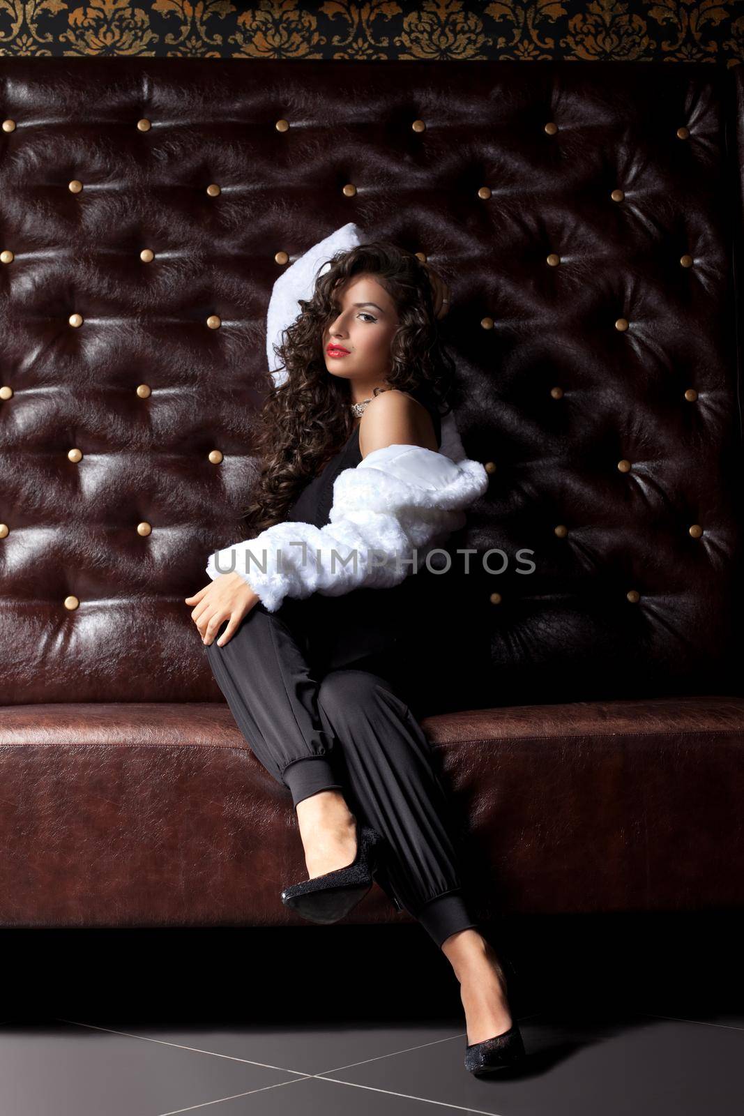 sexy young woman sit on leather sofa in fur coat in dark - look at camera