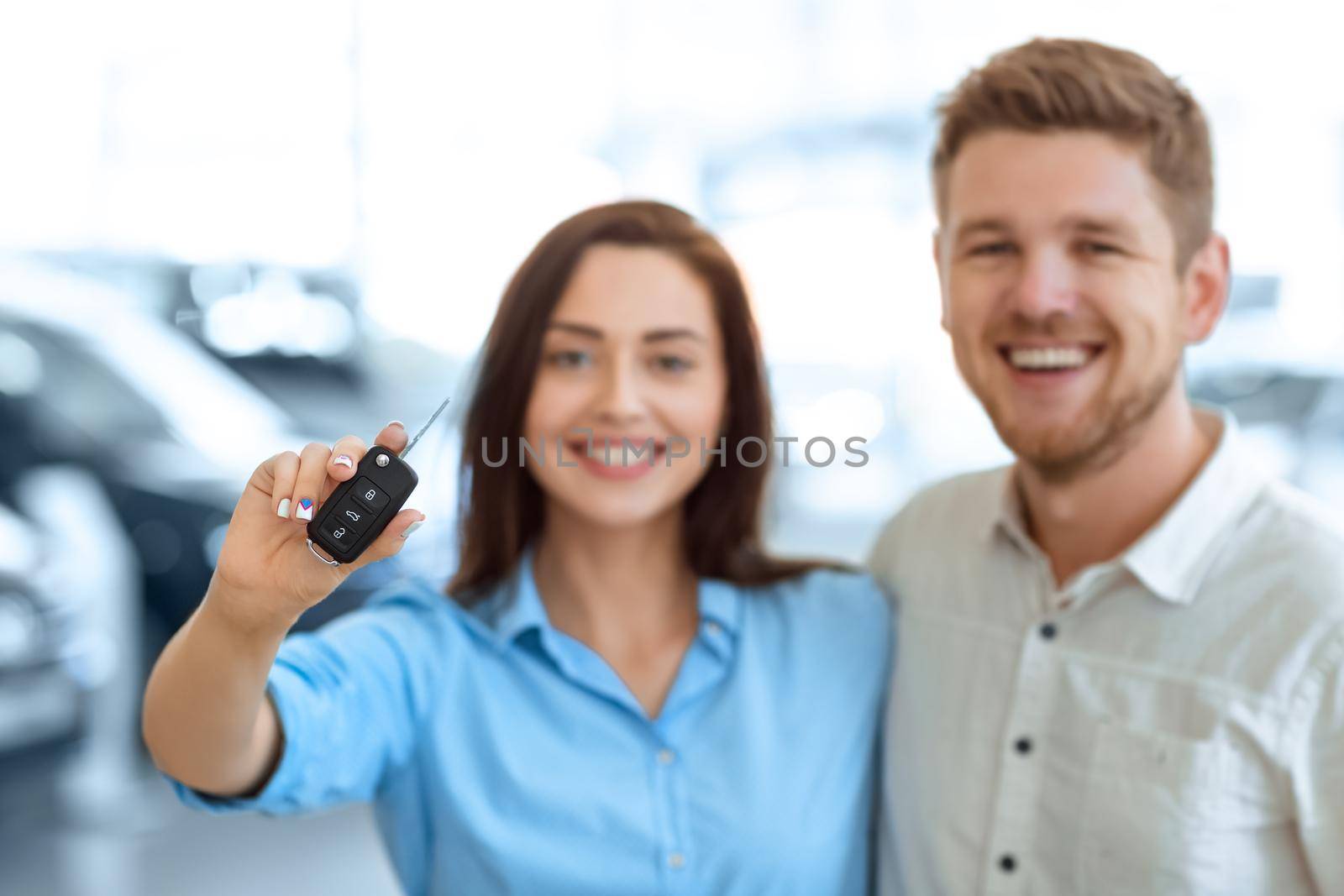 A key to the new chapter. Selective focus on a car key beautiful happy woman is holding hugging with her handsome husband in a car salon