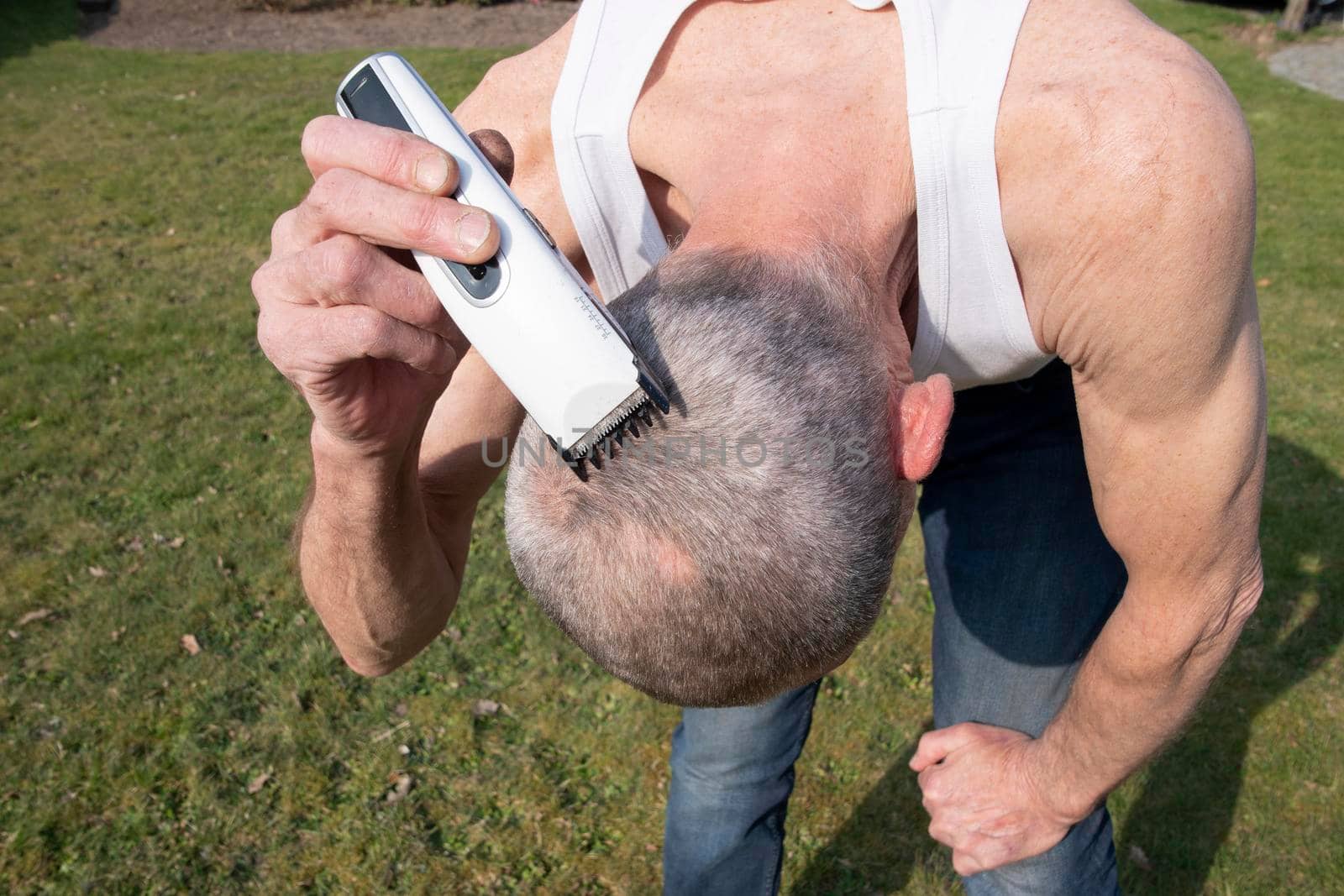 a gray-haired middle-aged man shaves his hair with a clipper in a garden on a green lawn, High quality photo