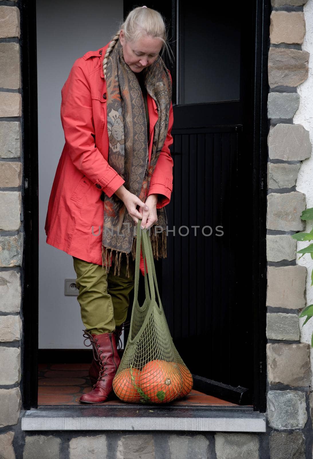 A middle-aged woman in a red cloak with a net of orange pumpkins opens the door by KaterinaDalemans