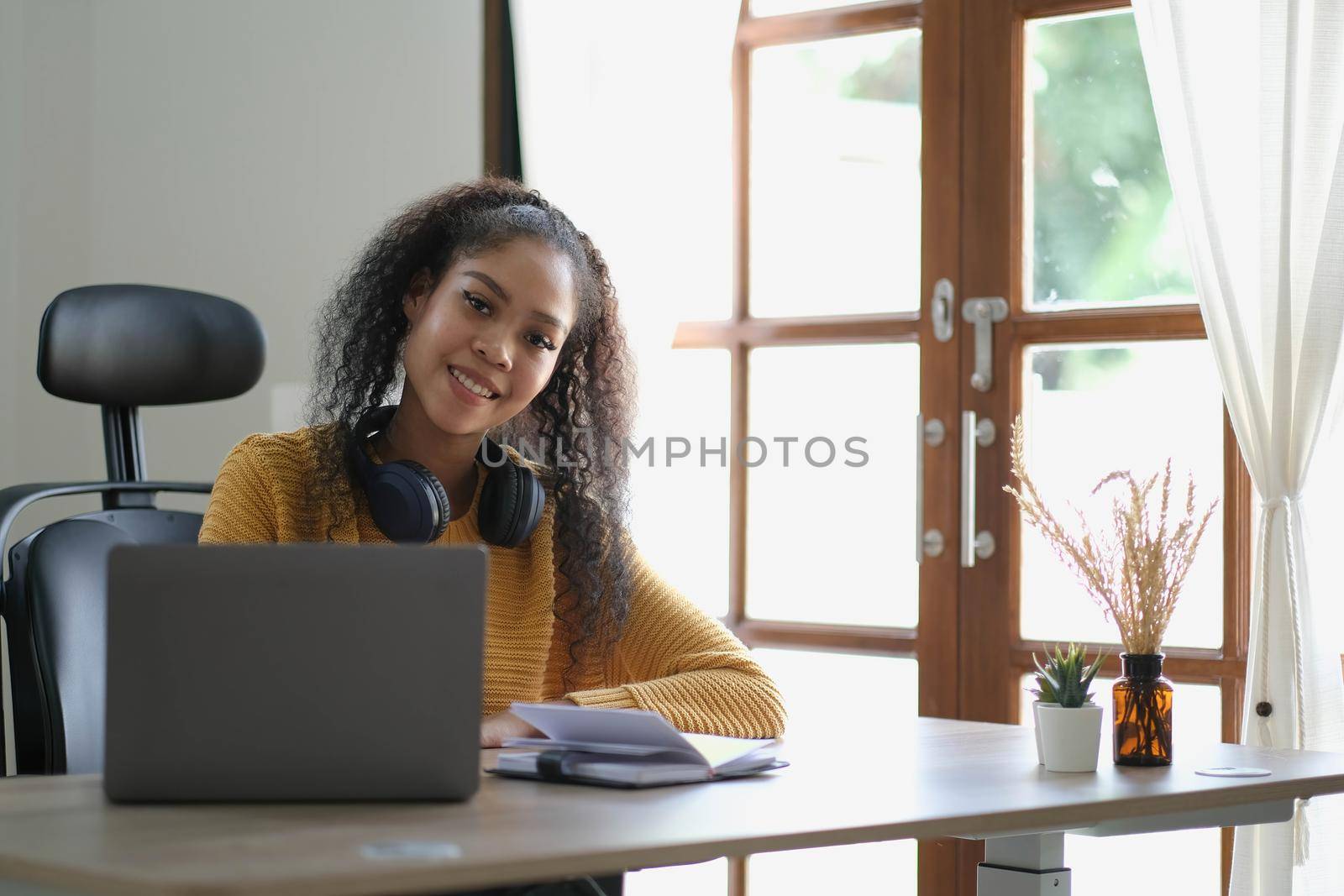 Smiling young African American woman in headphones distracted from computer work look in distance dreaming. Happy biracial female study online at home on laptop thinking or planning. Vision concept. by wichayada