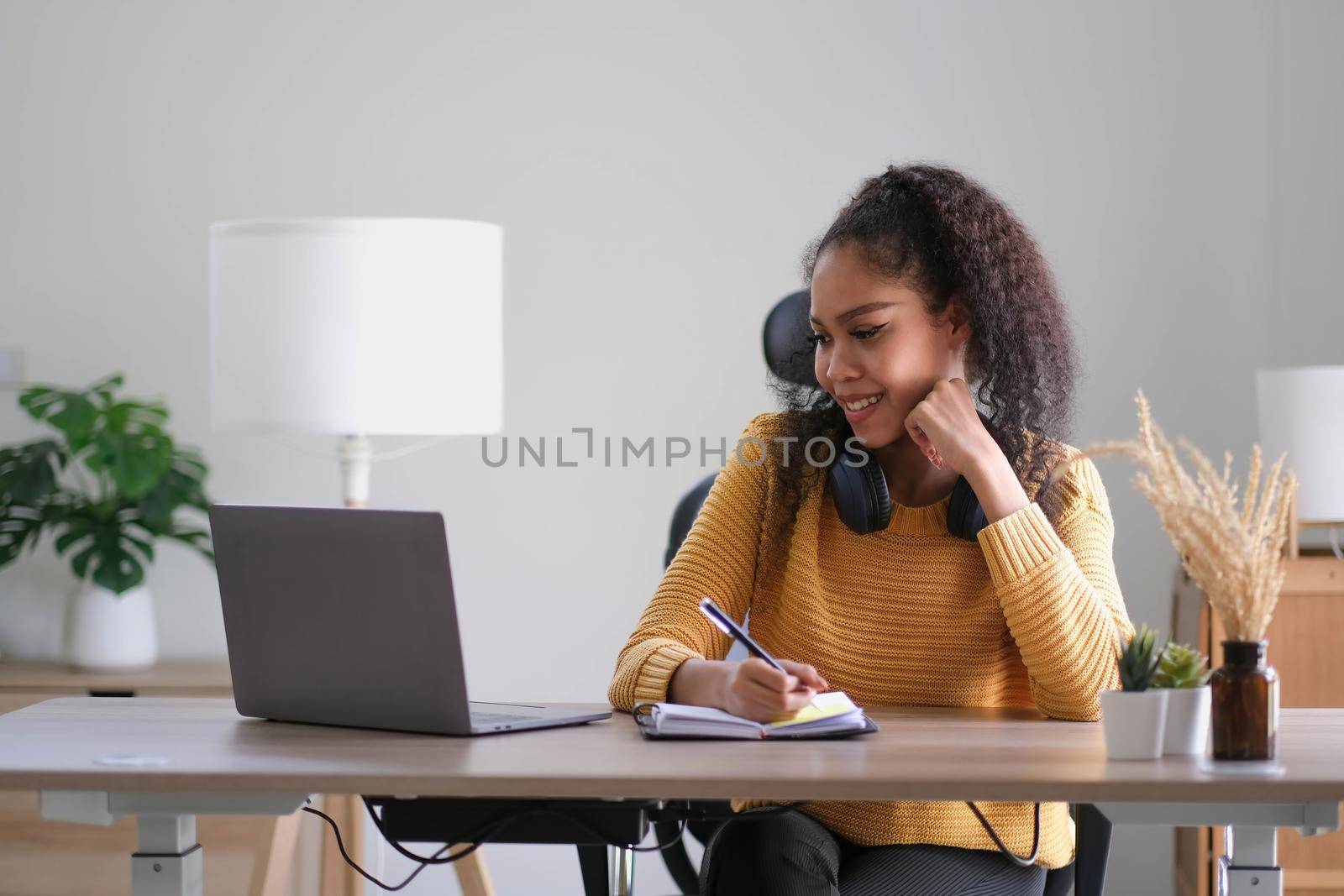 Young adult happy smiling Hispanic Asian student wearing headphones talking on online chat meeting using laptop in university campus or at virtual office. College female student learning remotely. by wichayada