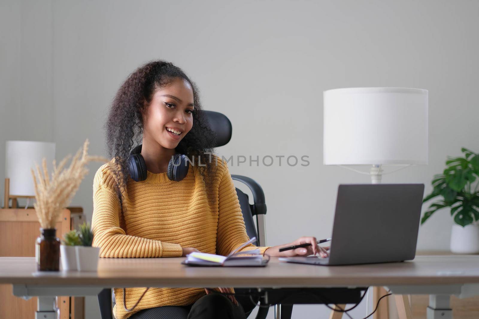 Young adult happy smiling Hispanic Asian student wearing headphones talking on online chat meeting using laptop in university campus or at virtual office. College female student learning remotely. by wichayada