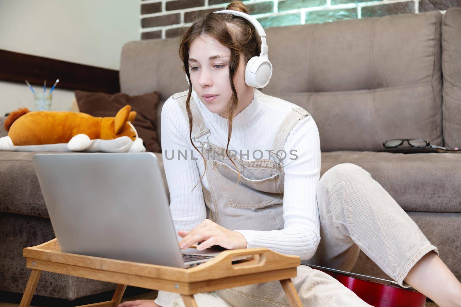 Smiling young woman with laptop watching content or learning while sitting at home in the living room on the floor in casual clothes and headphones by yanik88