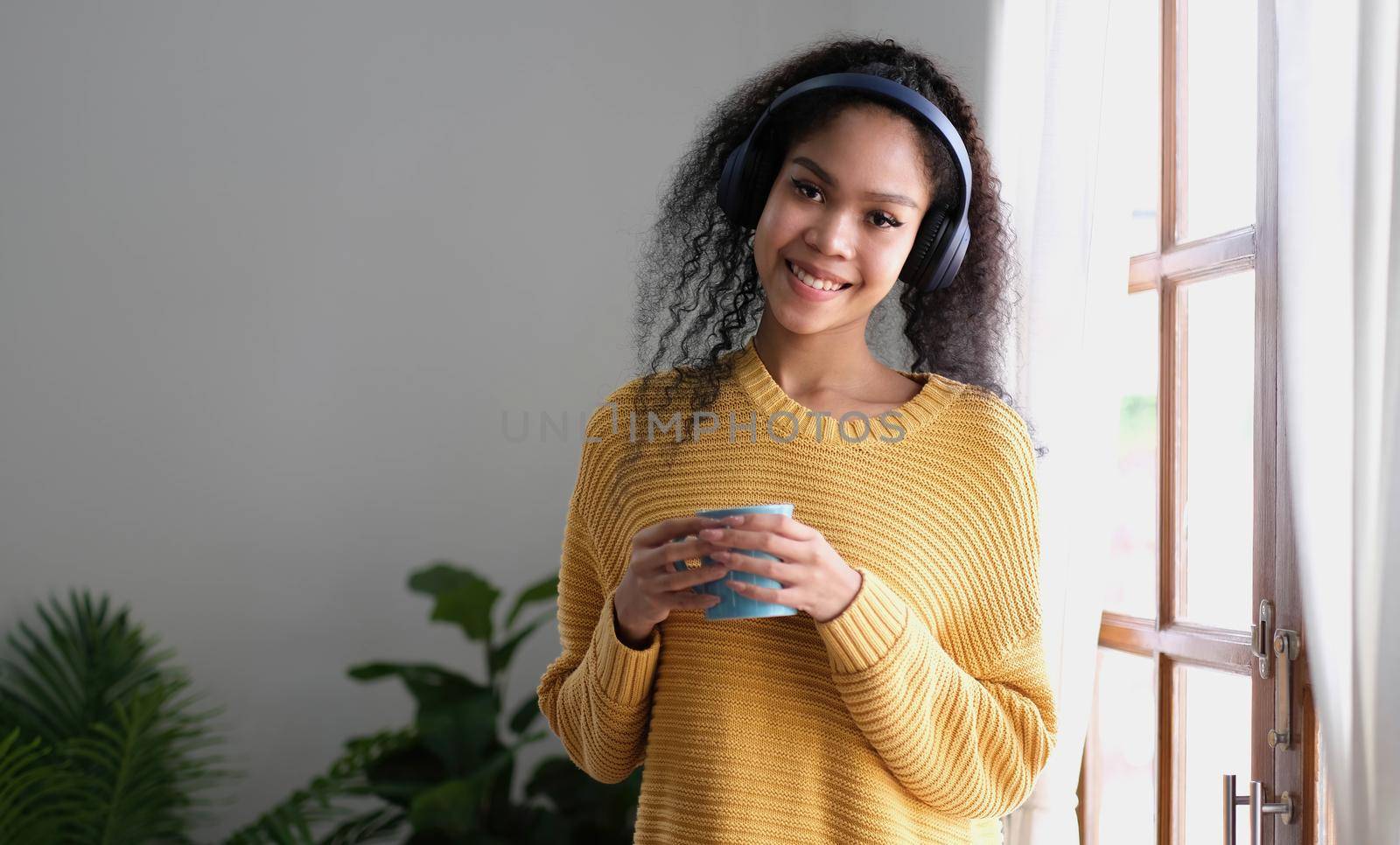 asian woman in headphones holding cup of coffee while standing near window at home by wichayada