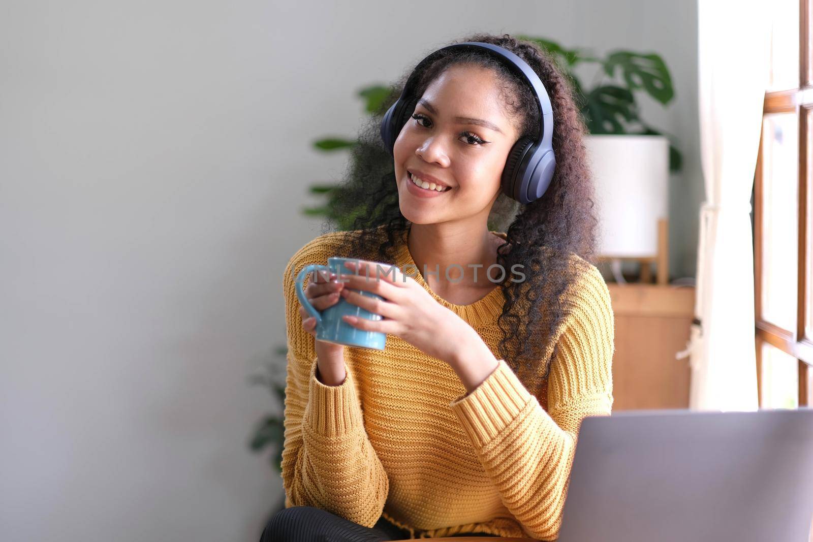 Smiling young asian woman using laptop web camera while holding a cup of coffee and wearing headphones in the living room at home..