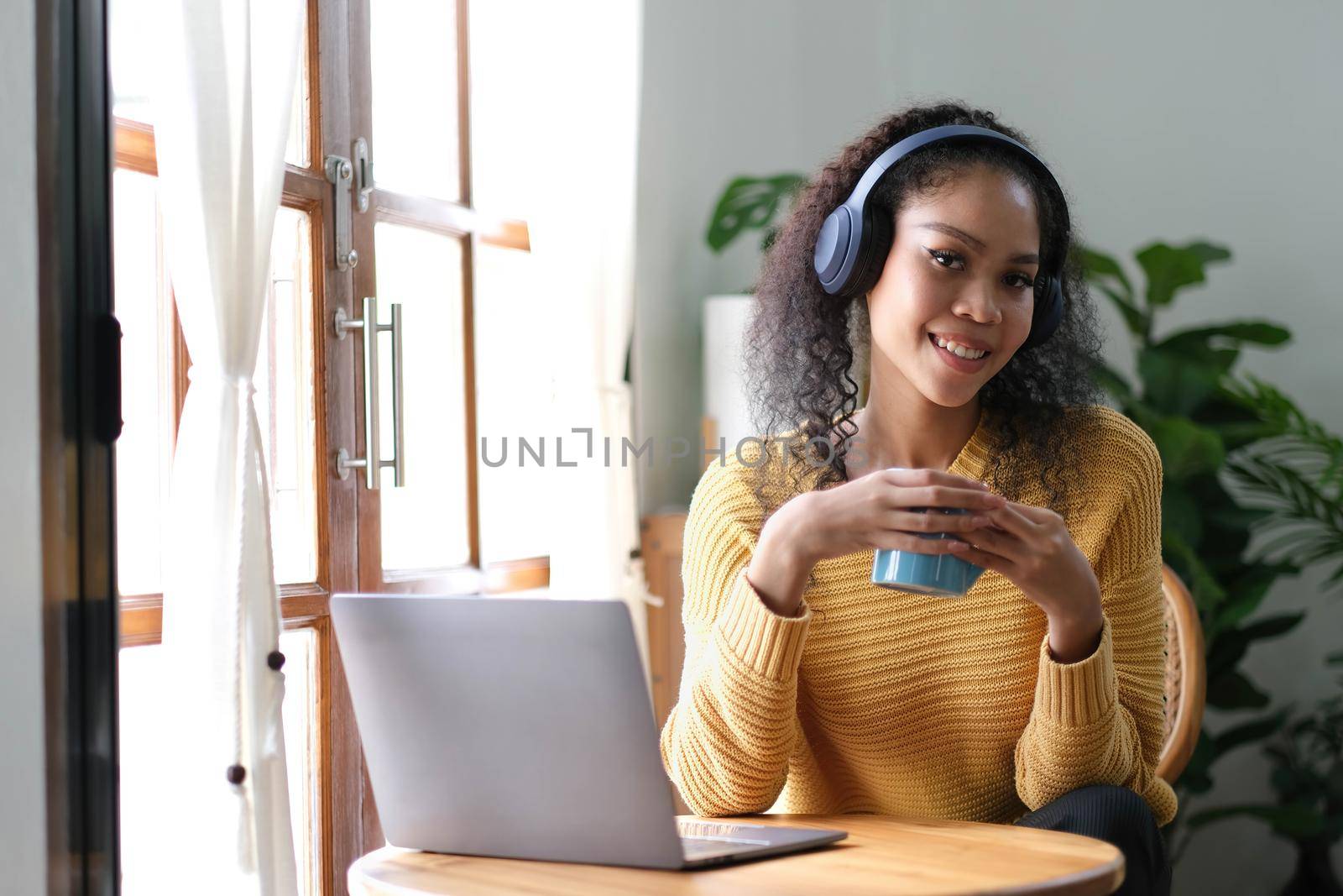 Smiling young asian woman using laptop web camera while holding a cup of coffee and wearing headphones in theliving room at home..