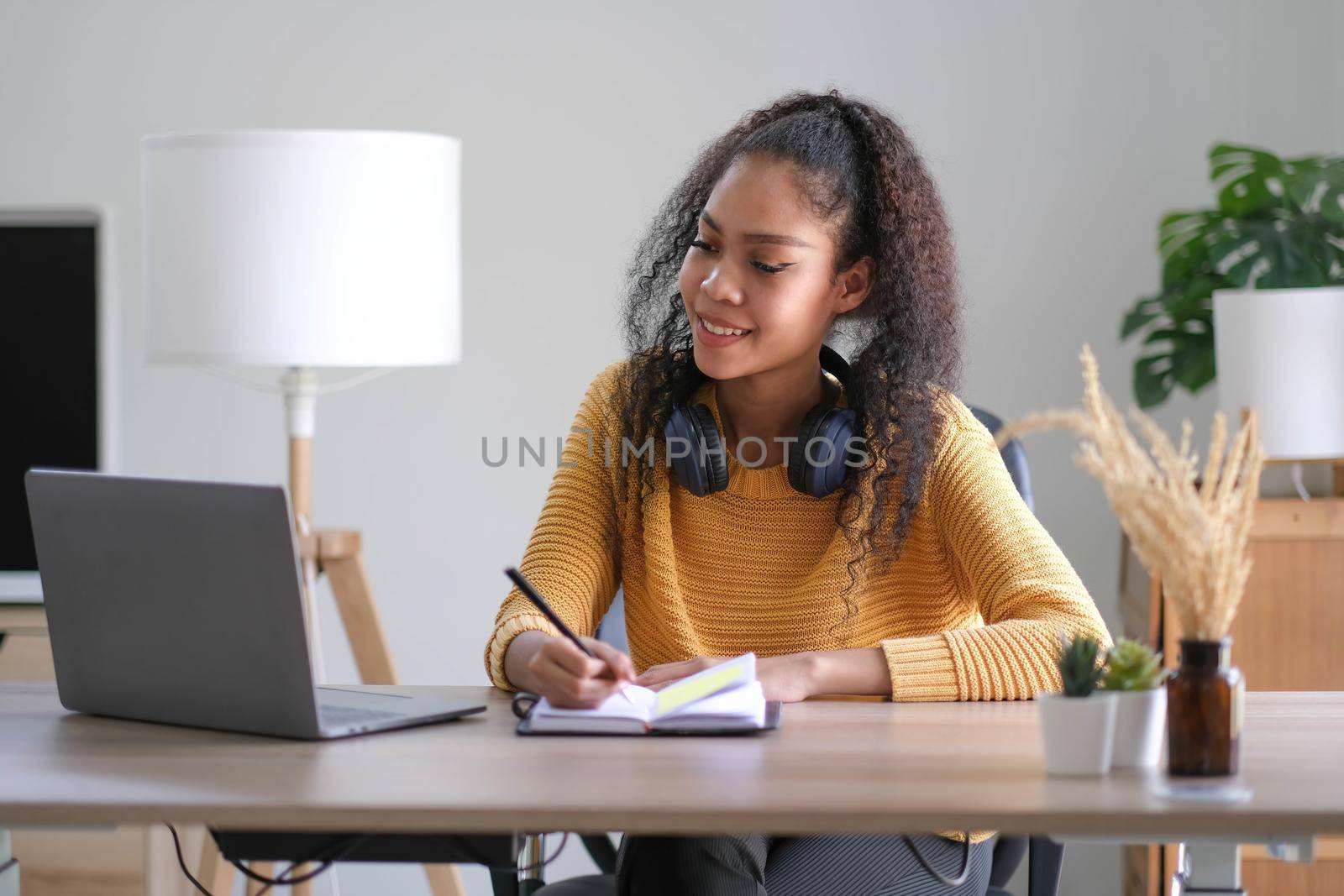 Smiling young African American woman in headphones distracted from computer work look in distance dreaming. Happy biracial female study online at home on laptop thinking or planning. Vision concept. by wichayada