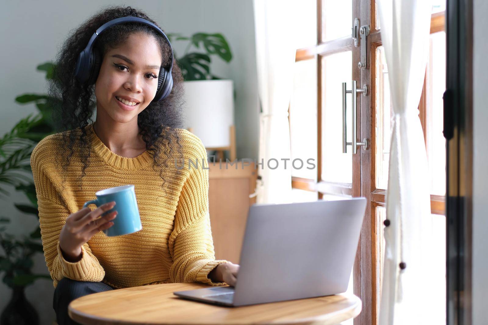 Smiling young asian woman using laptop web camera while holding a cup of coffee and wearing headphones in theliving room at home