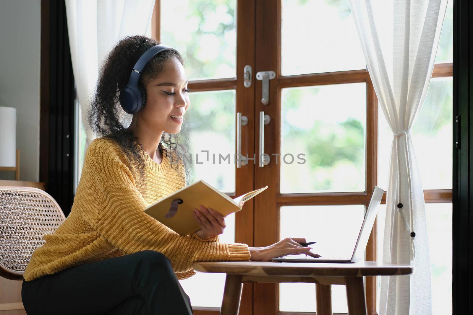 Smiling girl student wear wireless headphone study online with teacher, happy young asian woman learn language listen lecture watch webinar write notes look at laptop sit in cafe, distant education.