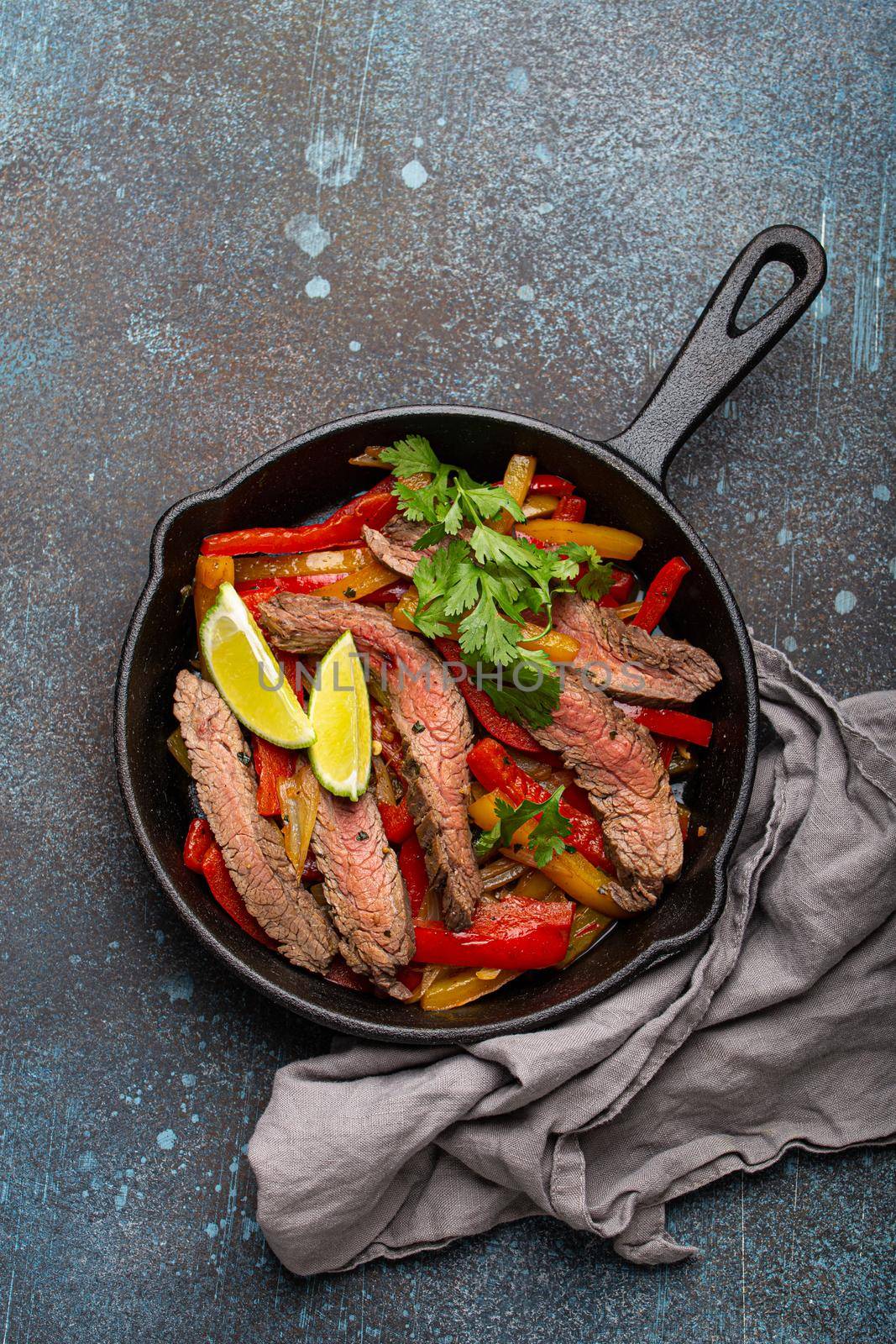 Traditional Mexican dish Beef fajitas with bell peppers in black cast iron pan on rustic stone background from above, American Mexican food