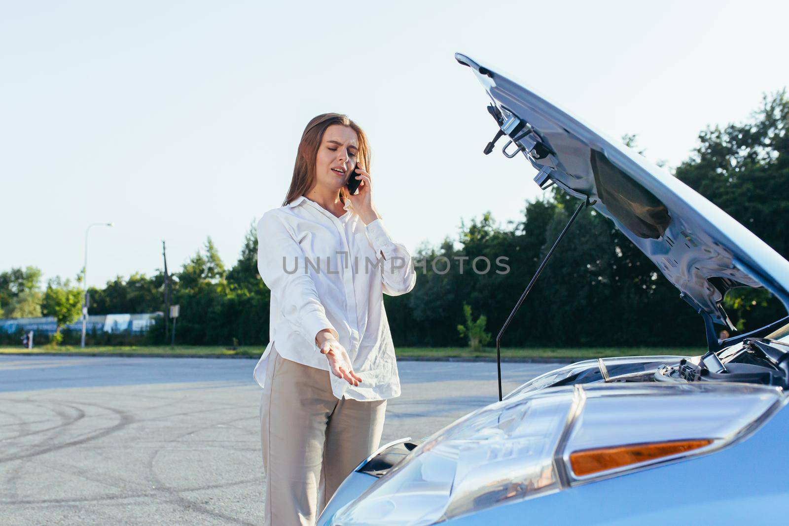 An annoyed and angry woman, near a broken car, tries to call an insurance agent and a repair crew. for car repair