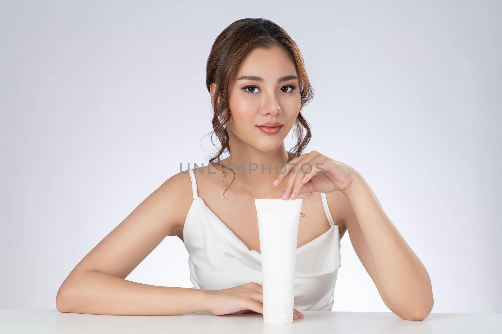 Gorgeous woman with makeup smiling holding mockup product for advertising text place, light grey background. Advertising concept for skin healthcare and beauty care products.