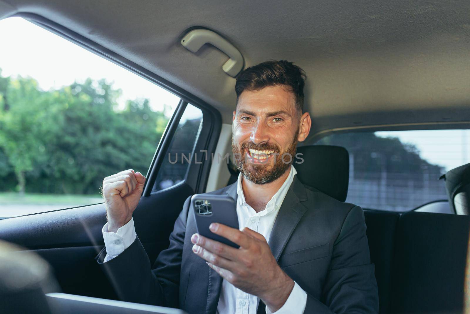 Happy and successful male businessman car passenger reads the news from the phone, rejoices in the victory and the news received