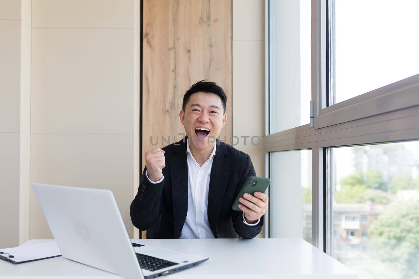 Happy young Asian business man in office, looking at camera, with emotions of winner or victory, financial promotion sports betting. The man exclaims happily while playing the game. Excited with joy, celebrating success