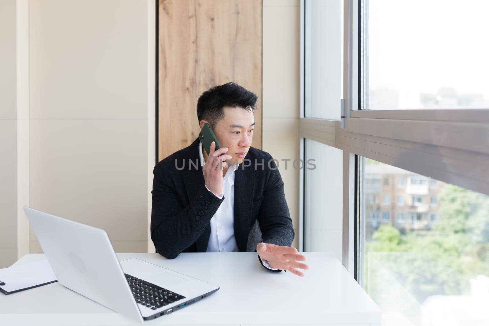 Serious asian business businessman uses cellphone to communicate with colleagues while sitting in modern office by voronaman