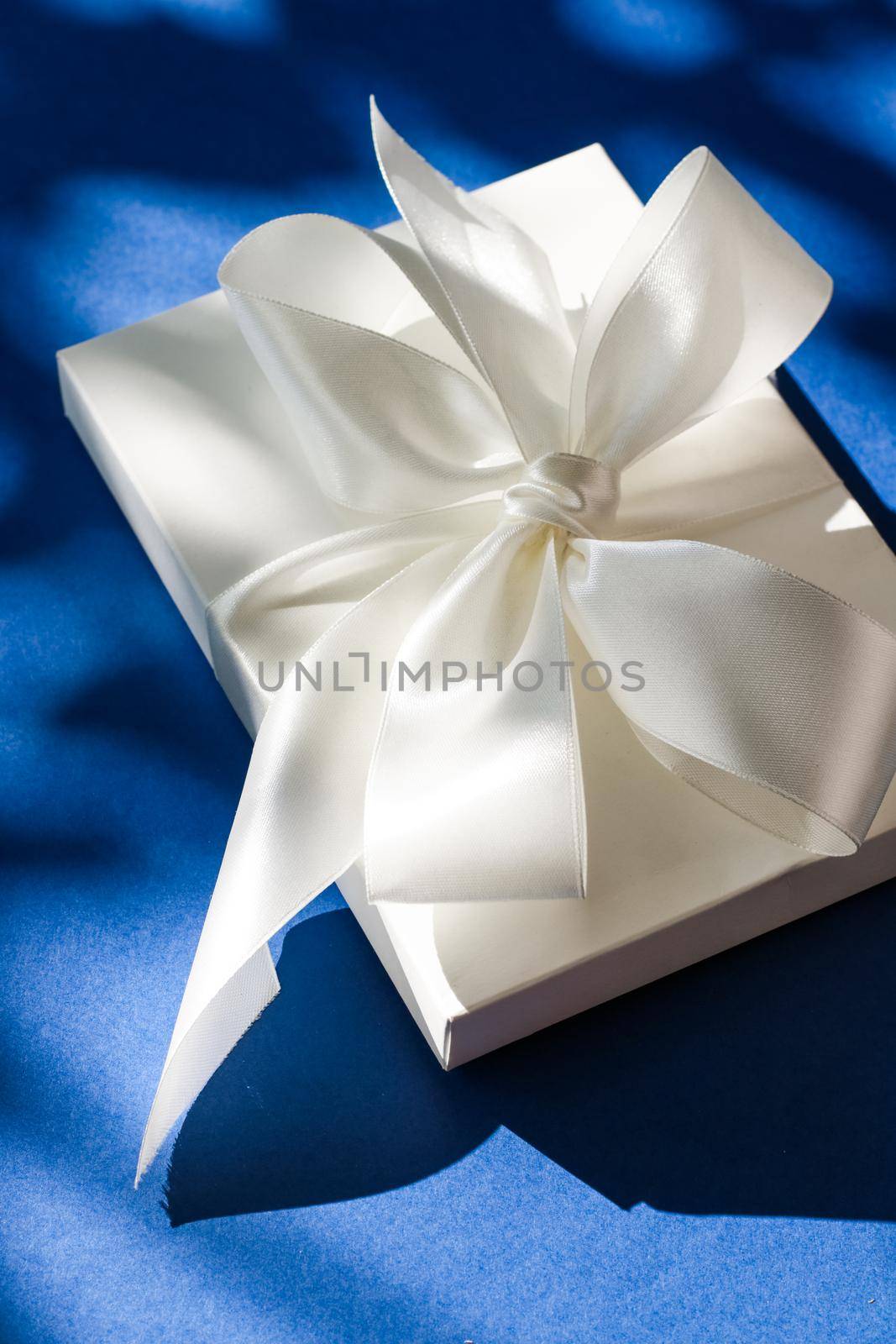 Luxury holiday white gift box with silk ribbon and bow on blue background, luxe wedding or birthday present by Anneleven