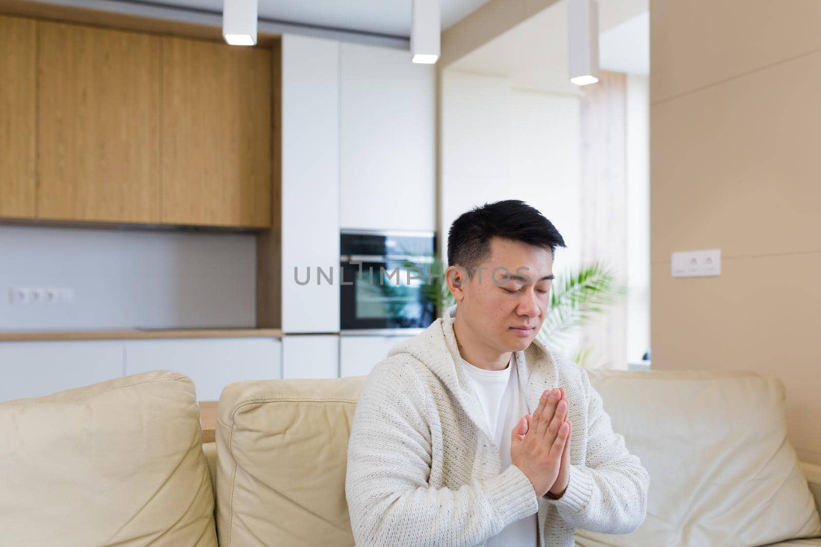 young asian man praying at home alone. Male folding pencil arms and meditating in apartment room. Christian religion traditional spiritual for their hope and believed god forgievness at house