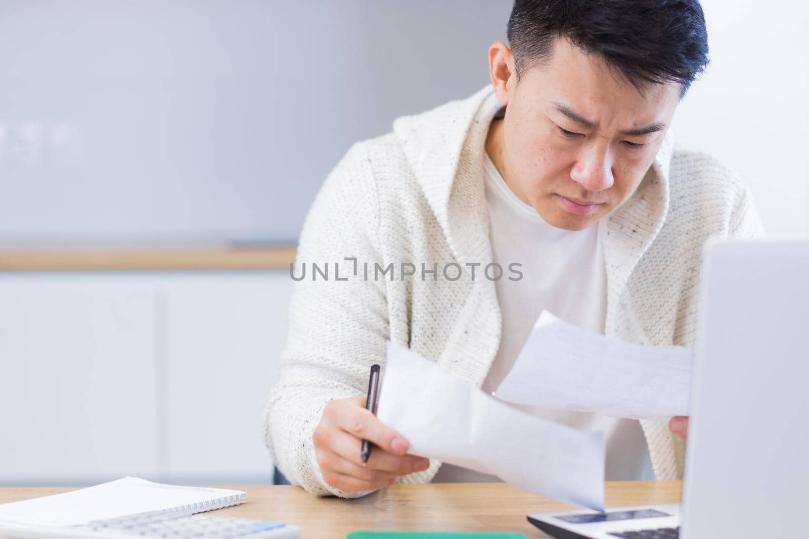 Young Asian man doing paperwork at home sitting at a desk, working on a laptop, focused on paying bills
